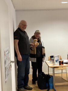 Hans und Siglinde stehen nebeneinander in einem Raum und halten gemeinsam einen Teddybären.