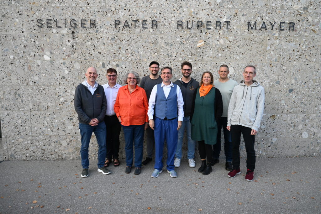 Gruppenfoto mit 9 Menschen. Sie stehen vor einer Wand aus Steinen mit dem Schriftzug "Seliger Pater Rupert Mayer".